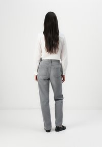 Una persona con cabello oscuro y largo lleva una camisa blanca y pantalones grises, vista desde atrás contra un fondo blanco minimalista.