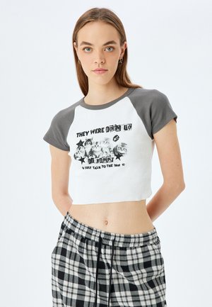T-shirt blanc court avec des manches grises, présentant un imprimé graphique de chats et du texte. Porté avec un pantalon à carreaux noir et blanc. Texture ribbed douce.