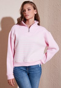 Felpa rosa con colletto a mezzo zip, caratterizzata da righe bianche orizzontali sul petto e polsini a costine, abbinata a jeans blu.