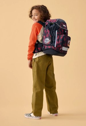 Lächendes Kind mit orange-beigem Oberteil, olivgrüner Hose, karierten Slip-on-Schuhen und einem großen rot-schwarz gemusterten Rucksack vor beigem Hintergrund.