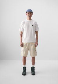 T-shirt blanche à manches courtes avec logo astérisque noir, shorts beiges avec détails sur les coutures latérales, et bottes de randonnée grises. Casquette bleue pour compléter le look.