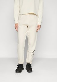 Cream-colored sweatpants made from soft fabric, featuring an elastic waistband and a graphic accent on the left thigh. Black athletic shoes.