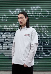 T-shirt blanc à manches courtes porté sur un t-shirt à manches longues rayé, avec un logo brodé. Fond : volet vert avec des graffitis.