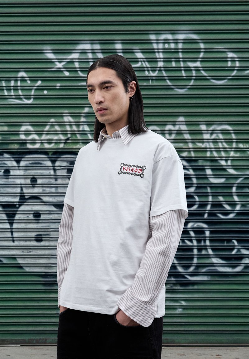 T-shirt blanc à manches courtes porté sur un t-shirt à manches longues rayé, avec un logo brodé. Fond : volet vert avec des graffitis.