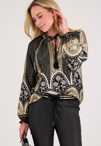 Zwart en beige gestippeld blouse met lange mouwen, strikjes bij de halslijn, bloemenpatronen en elastische manchetten. Gecombineerd met zwarte broek.