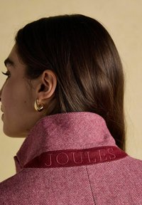 Žena s tmavo hnedými vlasmi nosí zlaté kruhové náušnice a štruktúrovanú ružovú bundu s vyšitým nápisom "JOULES" na vystúpenom golieri.