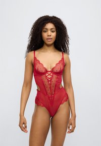 Κόκκινο κορμάκι από δαντέλα με διαφανή πάνελ, κυματοειδείς άκρες, ρυθμιζόμενους ιμάντες και σχέδιο σταυρωτού τύπου στο μπροστινό μέρος. Ανοιχτές πλευρές τονίζουν τη σιλουέτα.