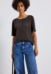 Donna con capelli ricci che indossa una maglietta a maniche corte marrone scuro infilata in un paio di jeans blu a vita alta, in piedi su uno sfondo chiaro e uniforme.
