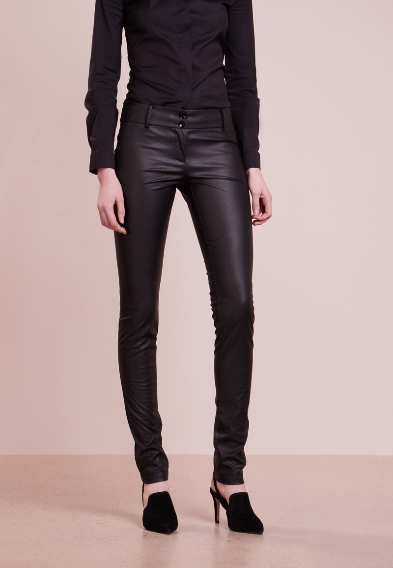 Pantalon skinny en similicuir noir à taille haute, avec une fermeture à deux boutons et une texture lisse et élégante, associé à des talons hauts noirs.