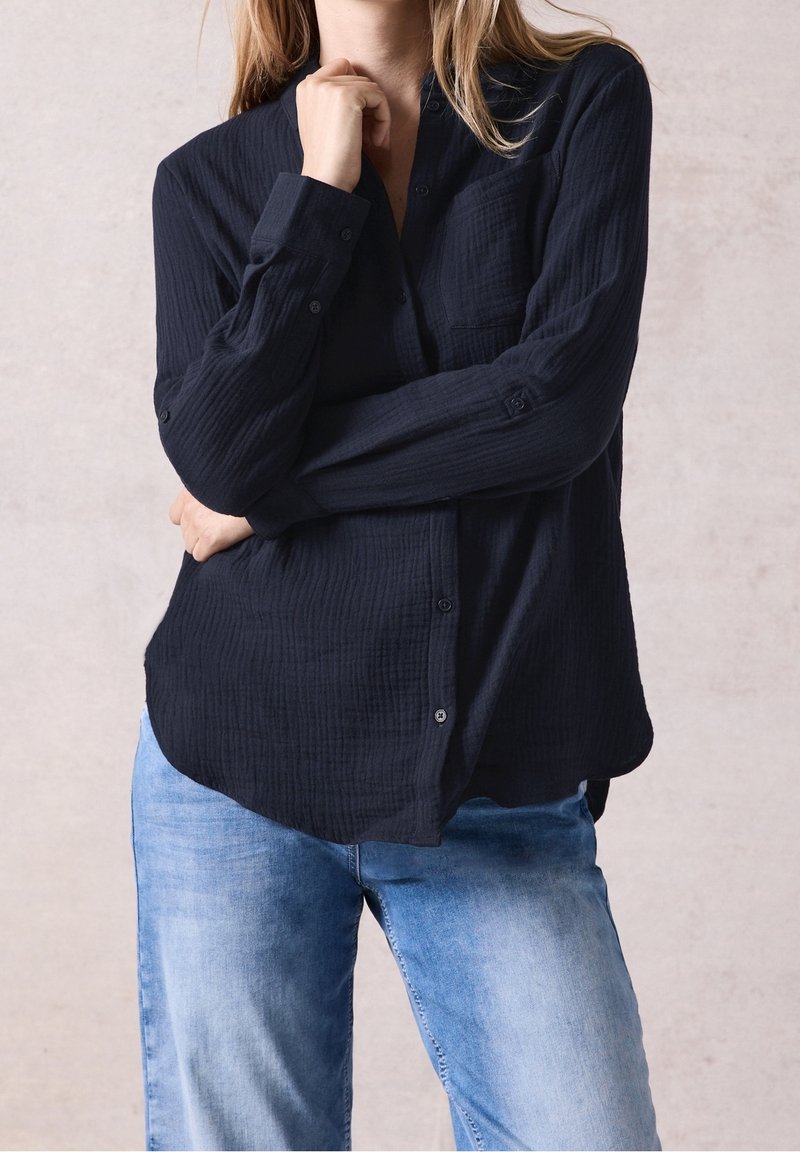 Personne portant une chemise noire à manches longues boutonnée et un jean bleu clair, avec un bras croisé et l'autre main touchant le menton.