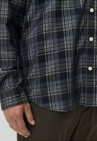 Marine geruite shirt met blauw en witte strepen, knopsluiting aan de voorkant, lange mouwen en een afgeronde zoom. De stof lijkt textuur te hebben met een zachte afwerking.