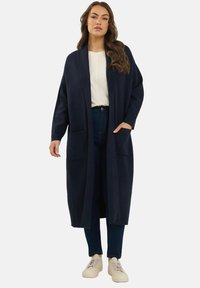 Cardigan lungo navy in tessuto morbido, design aperto davanti, con due tasche anteriori, indossato sopra una maglietta chiara e jeans scuri, con sneakers neutre.