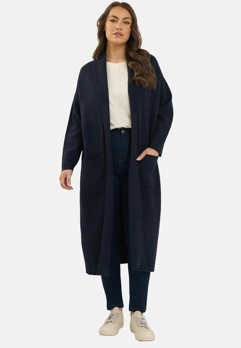 Cardigan lungo navy in tessuto morbido, design aperto davanti, con due tasche anteriori, indossato sopra una maglietta chiara e jeans scuri, con sneakers neutre.