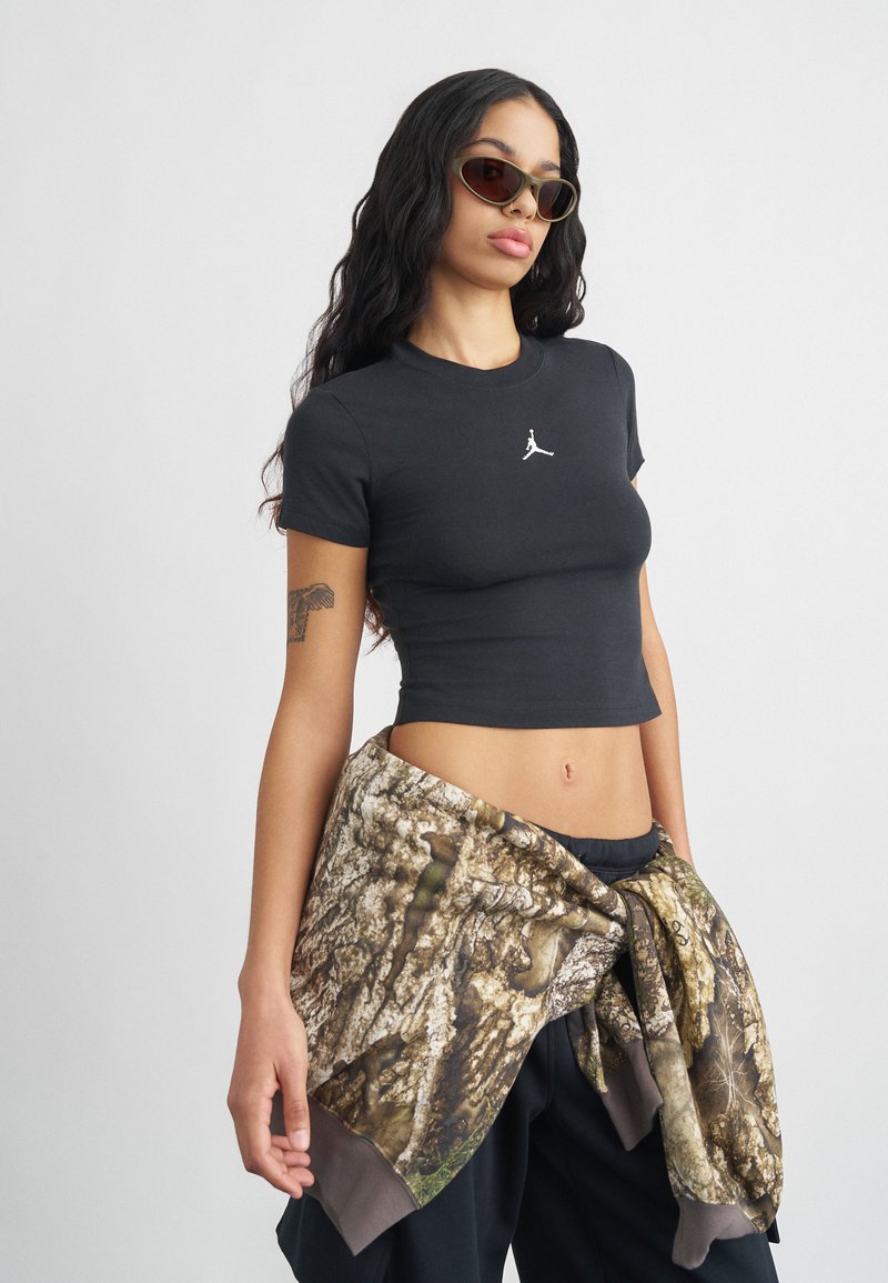 Νέο κορίτσι φοράει μαύρο crop top, γυαλιά ηλίου και στρατιωτική ζακέτα με καμουφλάζ δεμένη γύρω από τη μέση, στέκεται μπροστά σε απλό φόντο.