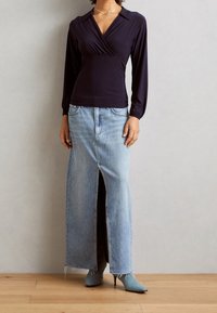 Marinblå långärmad topp med omslagsdesign; ljusblå, högmidjad denimkjol med mittslits; spetsiga blå stövlar; bakgrund av trägolv.