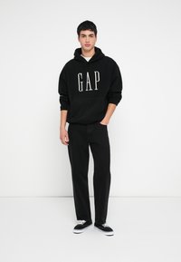 Mladý muž stojaci s jednou rukou v vrecku, oblečený v čiernej mikine GAP, čiernych džínsoch a čiernych teniskách s bielymi šnúrkami na bielom pozadí.