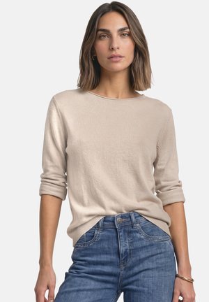 Femme aux cheveux bruns mi-longs portant un haut beige à manches longues et un jean bleu, debout devant un fond clair uni.