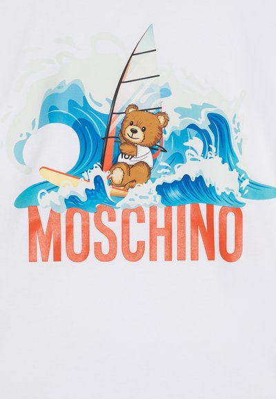 Bela majica s kratkimi rokavi, ki prikazuje risanega medveda na deski za surfanje med modrimi valovi, z izrazitimi rdečimi črkami "MOSCHINO" na dnu.