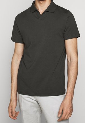 Mand iført en kortærmet mørkegrå polo t-shirt og lyse bukser, stående mod en ensfarvet baggrund.