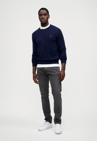 Suéter de malha azul-marinho com punhos canelados, sobre uma camisa branca com colarinho; jeans cinza ajustados e ténis brancos finalizam o visual.