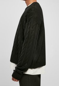 Suéter grueso de punto negro con puños y dobladillo acanalados, que presenta un escote redondo. Llevado sobre una camisa gris clara, mostrando una textura en capas.