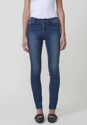 Femme portant un jean bleu foncé ajusté avec des poches avant et des chaussures noires à boucle, debout devant un fond clair uni.