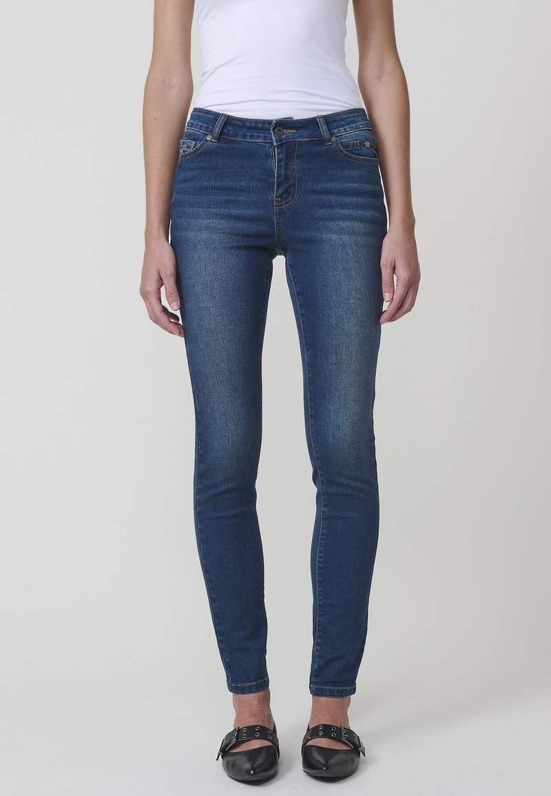 Femme portant un jean bleu foncé ajusté avec des poches avant et des chaussures noires à boucle, debout devant un fond clair uni.