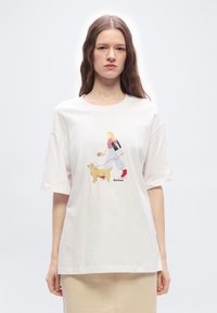 Γυναίκα με μακριά καστανά μαλλιά φοράει λευκό t-shirt με εικόνα μιας ξανθιάς γυναίκας που περπατάει ένα σκύλο, και οι δύο φορούν κόκκινα παπούτσια.