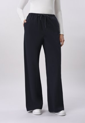 Person som har på seg løstsittende svarte bukser med snor og hvite joggesko, står med hendene i lommene mot en ensfarget bakgrunn.