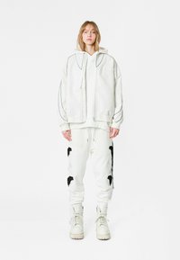 Sudadera blanca de gran tamaño con costuras negras en contraste, combinada con pantalones de chándal blancos que presentan bordados florales en negro, y botas blancas con cordones.