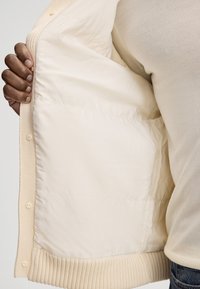 Veste beige avec ourlet et poignets côtelés, dotée d'un intérieur matelassé doux. Fermeture à un bouton sur le devant, tissu à texture lisse.