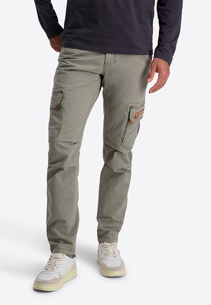 Man die lichtgroene cargobroek met meerdere zakken en witte sneakers draagt, staand tegen een effen achtergrond.