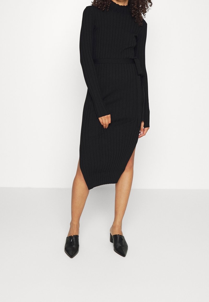 Femme portant une robe noire à manches longues en maille côtelée avec une ceinture à la taille et une fente latérale, associée à des mules à talons pointues noires.