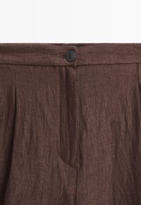 Brown linen trousers featuring a button closure, pleated front, and a textured finish. The waistband sits flat and smooth at the top.