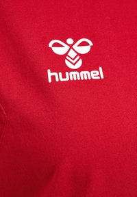 Rød t-shirt lavet af bomuld med en glat tekstur, med et hvidt Hummel-logo grafik på forsiden. Enkel design, korte ærmer.