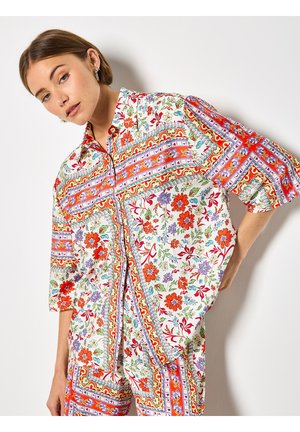 Jeune femme aux cheveux courts portant une chemise et un pantalon assortis aux motifs floraux et géométriques colorés, posant devant un mur blanc.