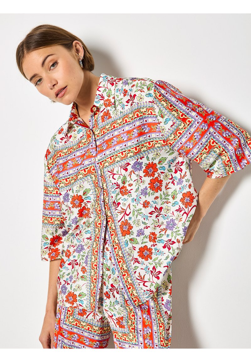 Jeune femme aux cheveux courts portant une chemise et un pantalon assortis aux motifs floraux et géométriques colorés, posant devant un mur blanc.