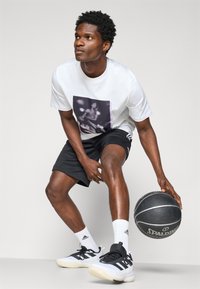 White T-shirt with a graphic print, black shorts, white and black basketball shoes, holding a black Spalding basketball, in a dynamic pose.