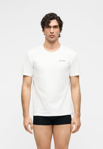 Νέος άνδρας με σκούρα σγουρά μαλλιά που φοράει λευκό T-shirt Moschino και μαύρα boxer, στέκεται μπροστά σε απλό ανοιχτό φόντο.