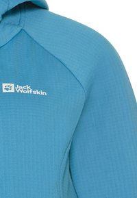 Haut de sport bleu avec un tissu texturé, comportant un logo blanc dans le coin supérieur gauche. Le design inclut une capuche et des coutures contrastées.