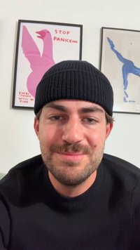 Căciulă neagră din tricot și pulover, ton de piele deschis, cu o barbă scurtă. Fundalul prezintă două afișe înrămate cu culori vibrante și text.