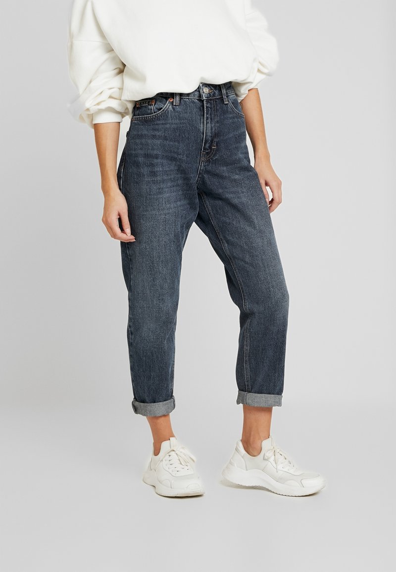 Person iført rullede mørkeblå jeans, hvide sneakers og en hvid langærmet top, stående mod en ensfarvet baggrund.