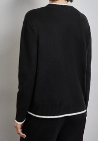 Osoba ubrana w czarny sweter z długim rękawem, z białym wykończeniem na kołnierzu, mankietach i dole, stojąca na tle jednolitej szarej ściany.