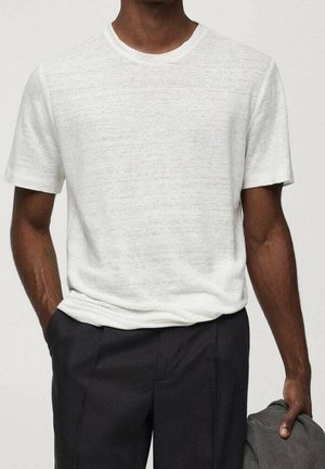 Mann trägt ein schlichtes weißes Kurzarm-T-Shirt und dunkle Hosen, hält ein gefaltetes graues Kleidungsstück in seiner rechten Hand.