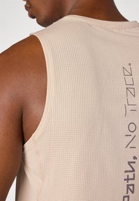 Beige ärmelloses Oberteil mit einem strukturierten Rautenmuster und kontrastierendem dunkeltext auf der Seite. Sanfter Stoff mit minimalen Nähten.