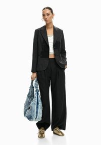 Blazer nero a righe sottili con design corto, abbinato a pantaloni neri larghi. Porta una borsa tote in denim azzurro chiaro e indossa sneakers oliva.