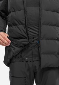 Schwarze gepolsterte Jacke mit einem Reißverschluss vorne und Druckknopfverschlüssen, die ein gefüttertes Innenfutter und verstellbare Manschetten zeigt, kombiniert mit passenden Hosen.