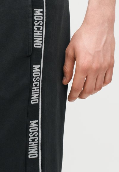 Κοντινό πλάνο ενός χεριού που ακουμπά δίπλα σε μαύρο παντελόνι με κάθετη λωρίδα λογότυπου Moschino στο πλάι.