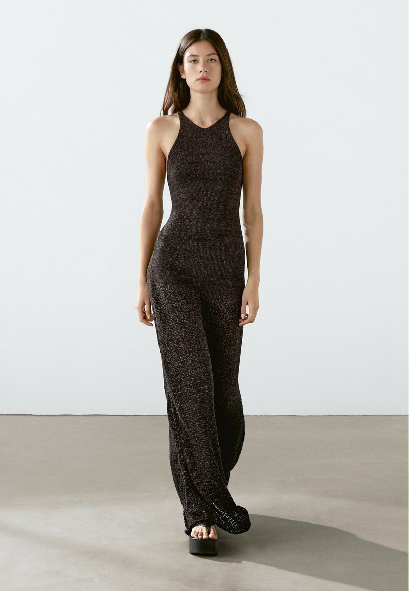 Jumpsuit nero in maglia con corpetto fitted, pantaloni larghi e collo all'americani. Il materiale ha una finitura testurizzata. La modella indossa sandali platform neri chunky.