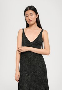 Vestido preto sem mangas com um decote em V profundo, apresentando uma textura pregueada e fios metálicos por todo o tecido para um efeito cintilante.